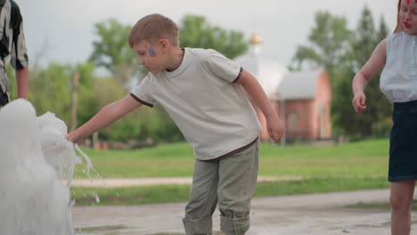 playful kids running near a water jet in park, splashing and laughing, boy reaching toward the spray while a woman walks by, summer outdoor scene showing movement, joy, interaction, blurred greenery