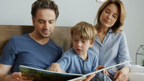 young mother and father sitting on the bed in the morning with their little son and reading an interesting book