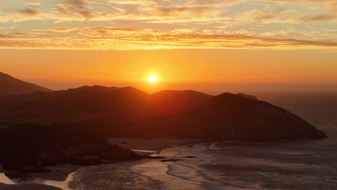 Uma ampla vista aérea da costa da Ilha Sul da Nova Zelândia no auge do pôr do sol, capturando o intenso brilho dourado sobre as montanhas e as ondas rítmicas quebrando na costa.