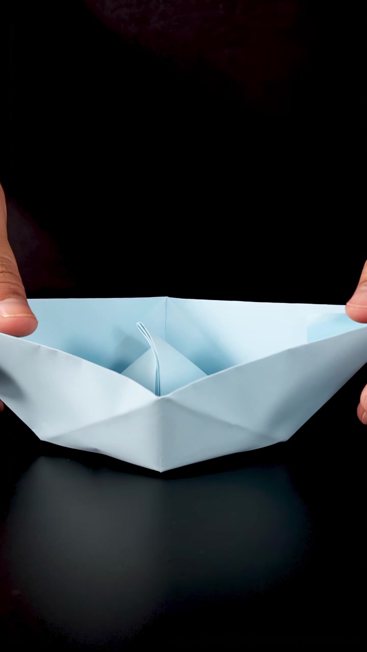 Person folder dygtigt blåt papir til en origami-båd, nærbillede, under blød studiebelysning, statisk kamera.