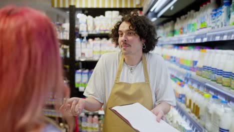 Over-the-shoulder-a-happy-guy-with-curly-hair-holds-a-tablet-in-his-hands-and-does-an-inventory-while-communicating-with-customers-in-the-dairy-department-of-a-supermarket