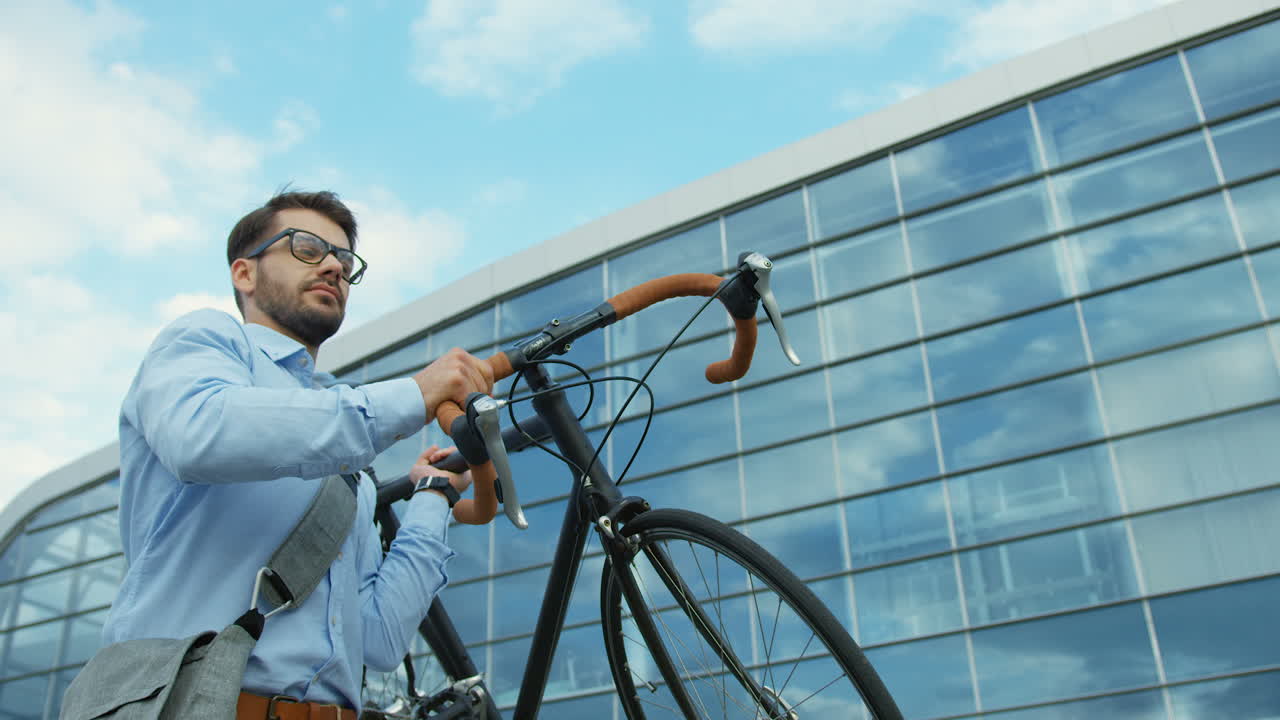 hombre guapo con gafas y estilo informal llevando su bicicleta, luego bajándola, sentándose en ella y montando cerca de un edificio de cristal