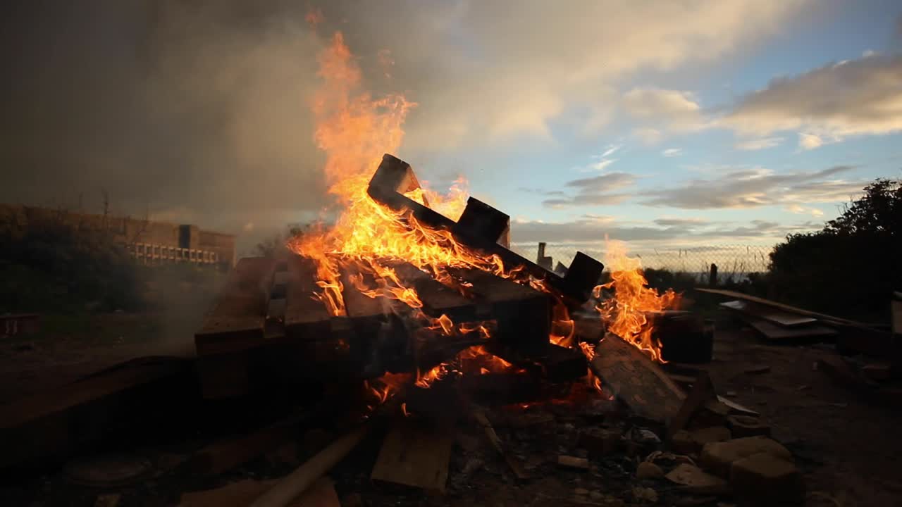 Large fire burning in a backyard. Crates and wooden rubble are visible in a pit. Smoke is coming towards the camera. Sun is setting and cloudy sky look beautiful. Shot in wide angle. Footage in HD
