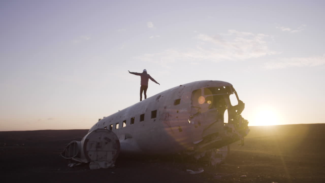 Giovane viaggiatore che cammina su un aereo schiantato e abbandonato su una spiaggia di sabbia nera al tramonto.
