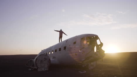 Joven-Viajero-Caminando-En-Un-Avión-Estrellado-Y-Abandonado-En-Una-Playa-De-Arena-Negra-Al-Atardecer