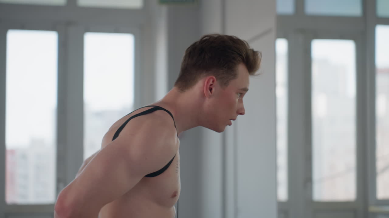 young man wearing resistance rope over shoulders bends forward with focused expression while looking at mirror in bright fitness room, preparing for workout