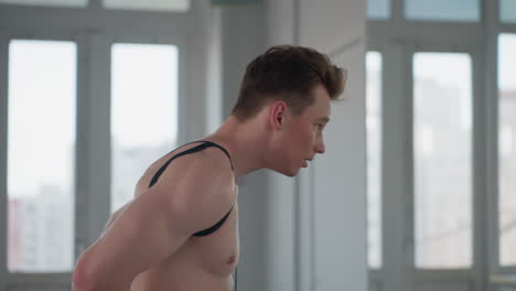 young man wearing resistance rope over shoulders bends forward with focused expression while looking at mirror in bright fitness room, preparing for workout