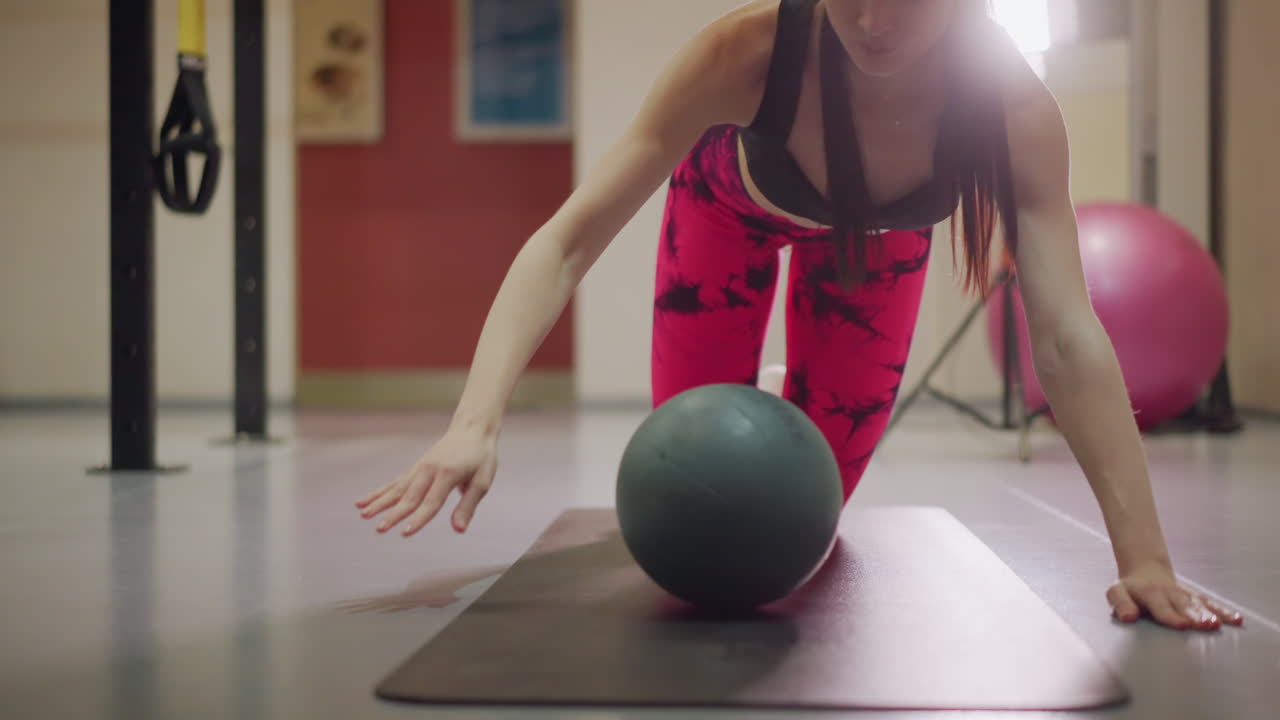 mujer asiática arrodillada con balón medicinal sobre colchoneta en un gimnasio luminoso, realizando un alcance con un brazo y una variación de plancha, ambiente de entrenador concentrado con luces de estudio, entrenamiento funcional para la estabilidad y el hombro