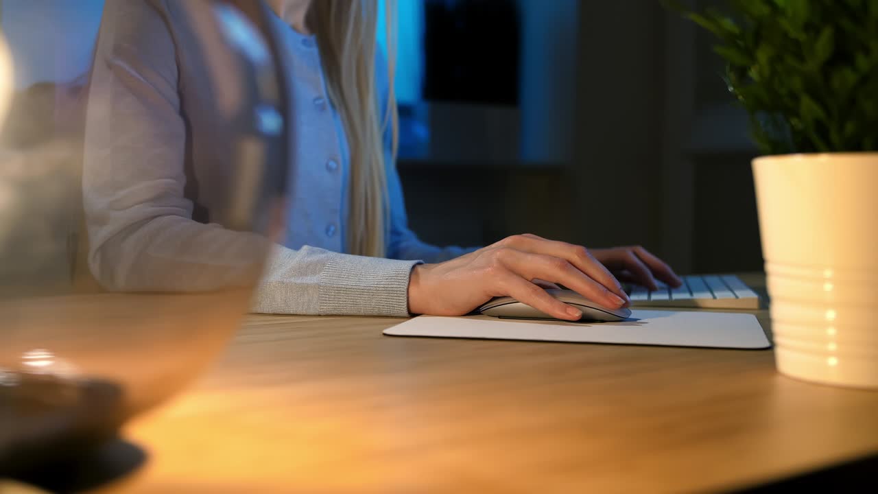 Vrouw werkt 's nachts op de computer. Close-up van de handen van een vrouw in comfortabele huiskleding, zittend aan een houten bureau, verlicht door een lamp, en gebruikt een computermuis en toetsenbord in een donkere kamer 's nachts.