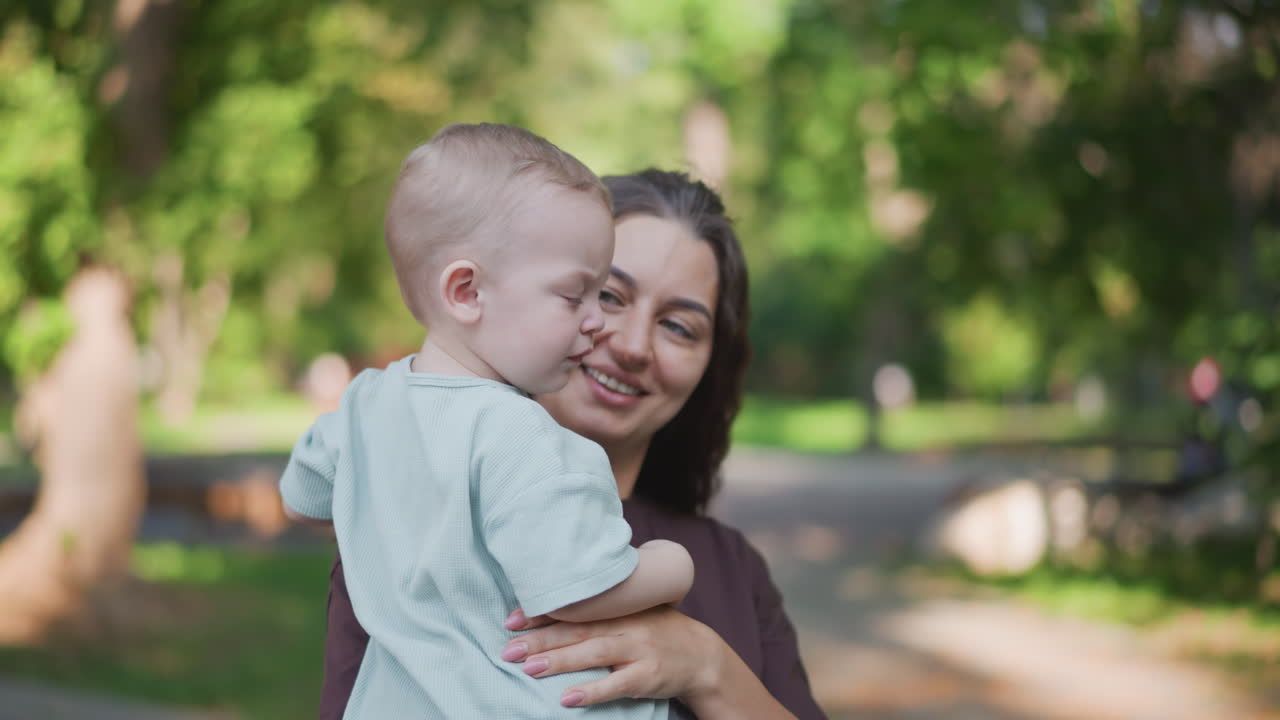 Madre latina che bacia il bambino piccolo al parco, un tenero e stretto abbraccio con un sorriso caloroso tra sentieri alberati, un momento affettuoso all'aperto che trasmette cura, protezione e la serenità di uno stile di vita familiare.