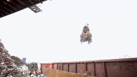 Crane-unloading-scrap-metal-into-a-dumpster-at-a-recycling-plant,-overcast-day