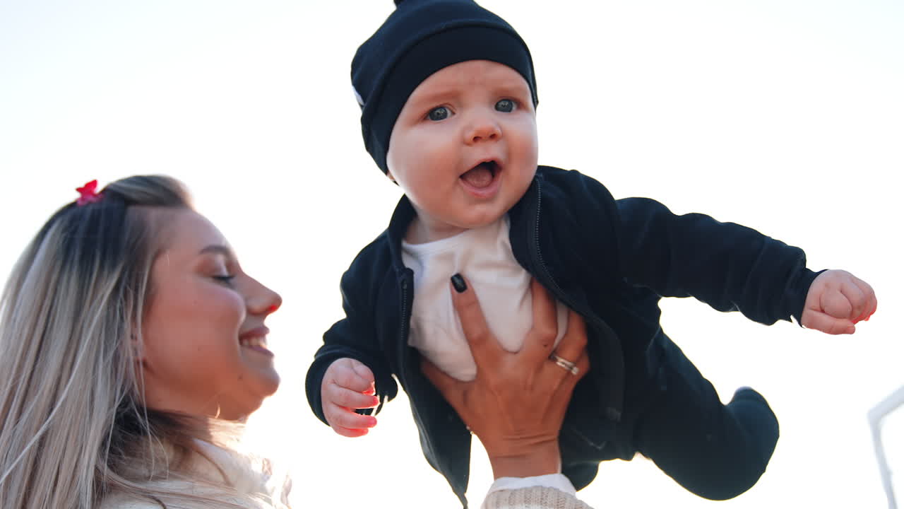 Happy long-haired mom holding her baby boy outdoors. Mother is tossing the kid looking into camera. Low angle view.