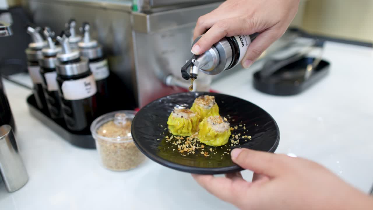 Une personne verse de la sauce foncée sur des raviolis shumai jaunes sur une assiette noire à Bangkok.