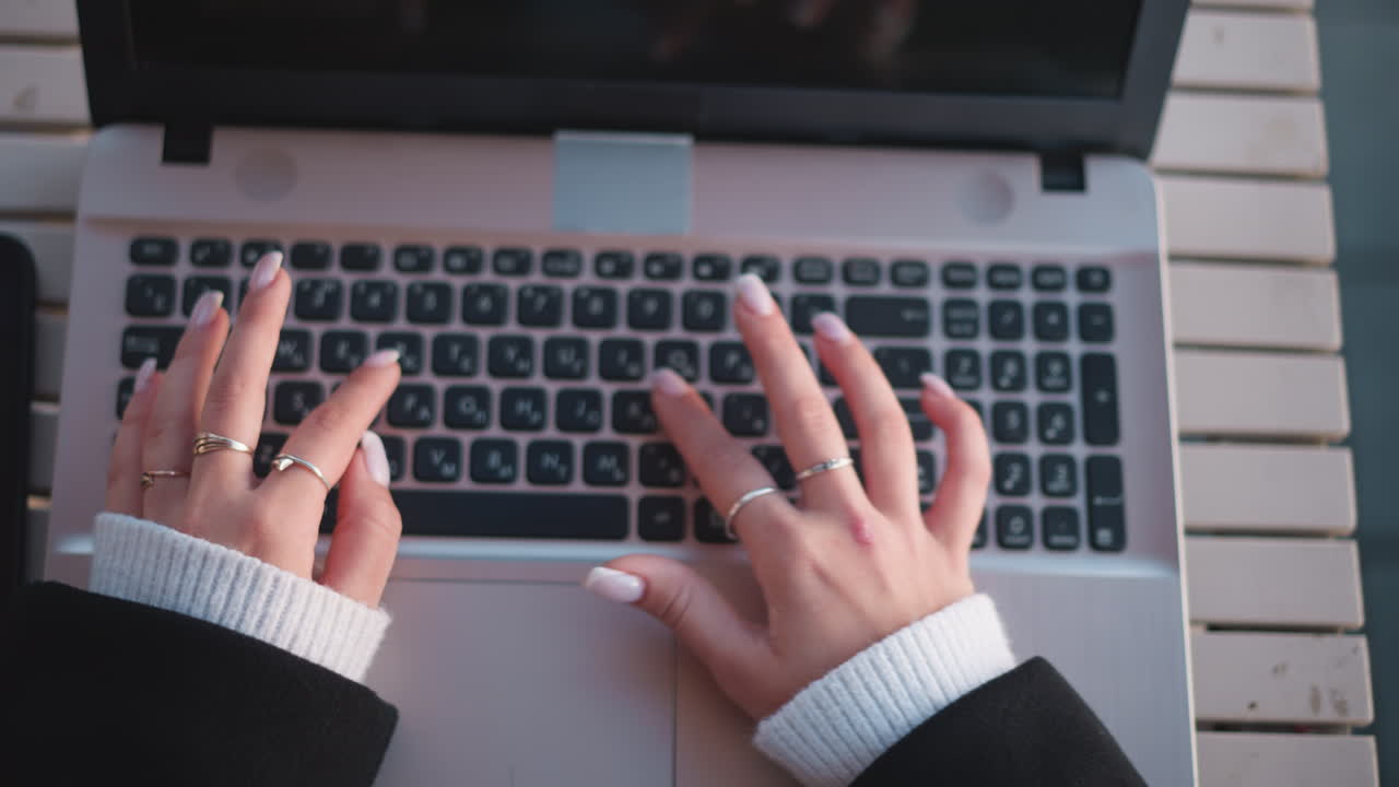 primer plano de manos con uñas pulidas escribiendo en un teclado de portátil, adornado con elegantes anillos, cómodas mangas de suéter y un teléfono inteligente en una mesa de madera