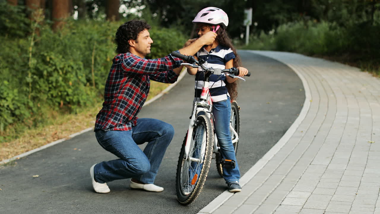 Closeup. Portrait of a pretty girl and her father near the bike. Dad wears a helmet on the girl's head. Smiling. Blurred background