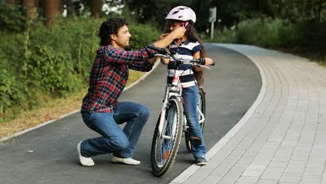 Closeup.-Portrait-of-a-pretty-girl-and-her-father-near-the-bike.-Dad-wears-a-helmet-on-the-girl's-head.-Smiling.-Blurred-background