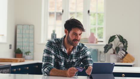 Celebrating-Man-With-Credit-Card-Using-Digital-Tablet-At-Home-To-Book-Holiday-Or-Shop