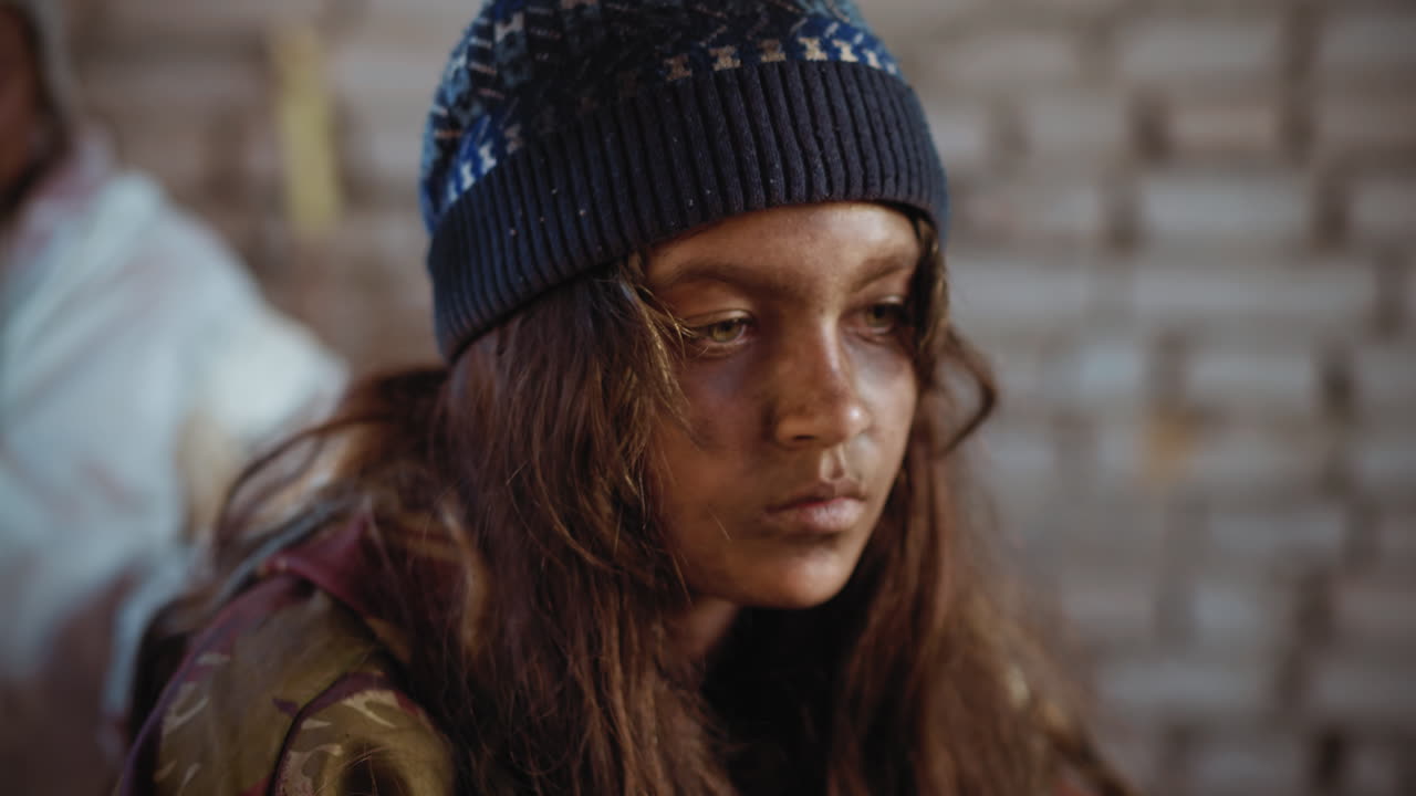 Sad haggard girl with dirty face and tired eyes sits in worn-out clothes and winter hat inside cold abandoned building, lost in thought while blurry person remains in background unnoticed