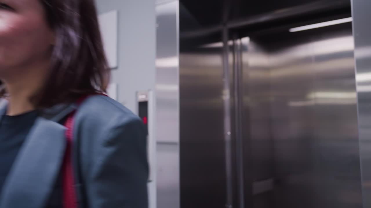 Businesswoman walking out of elevator in modern office, smiling confidently. Dressed in formal business suit, carrying a red handbag. Clean, sleek elevator interior and professional environment