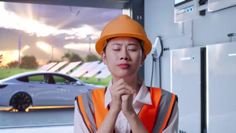 close up of asian female engineer with safety helmet pray for something with home energy storage system in a modern garage