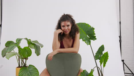 dark curly haired and tanned model posing on a chair with a red bikini suit, surrounded by plants, shot in a photo studio