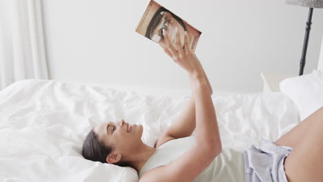 Happy-biracial-woman-lying-on-bed-reading-book-at-home,-slow-motion