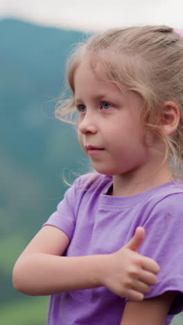 positive little child with blonde hair shows thumb-up gesture to parents over green valley against misty mountains on overcast day closeup slow motion