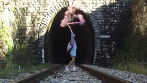 person holding pink smoke in tunnel on railroad tracks