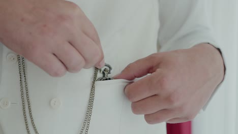 close-up of a groom adjusting a pocket square in his white suit, with a silver chain accessory
