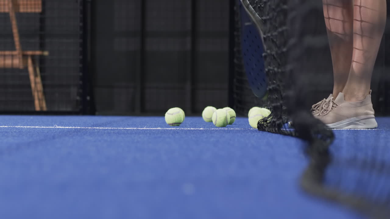 White woman stepping behind net on padel court clearing yellow balls using racket, copy space