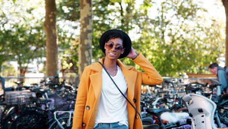 fashionable young black woman wearing a hat, sunglasses, blue jeans, an unbuttoned yellow pea coat and a crossbody handbag walking amongst parked bicycles, smiling to camera