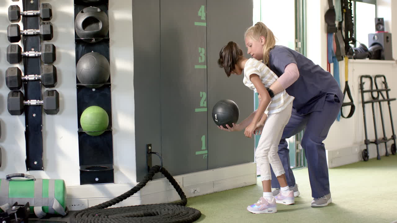 girl with cerebral palsy in rehabilitation, therapist assisting with medicine ball exercise in gym