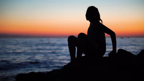 una joven sentada en las rocas de la costa del mar mientras hace su ejercicio matutino diario con un hermoso amanecer en el fondo - plano general