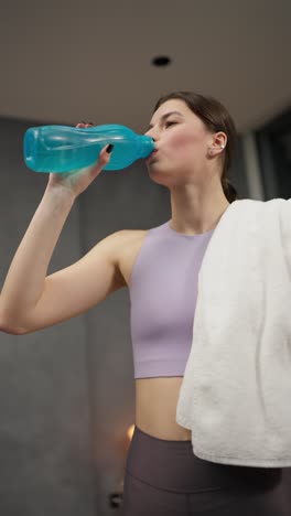 vertical video of a confident athletic brunette girl in a purple top drinks water from a blue and sports bottle after her exercise session in a modern apartment at home