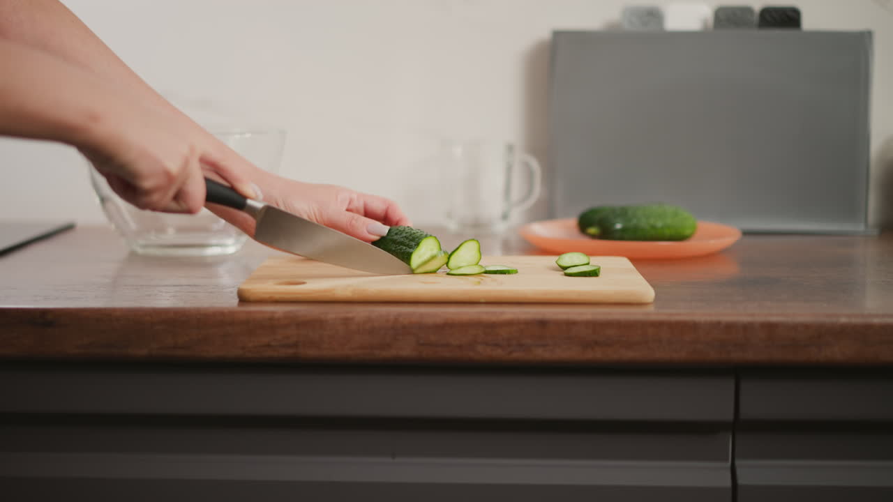 Close up of hand slicing fresh cucumber on wooden cutting board using sharp knife, with glass bowl and orange plate in background on modern countertop inside bright, organized home kitchen