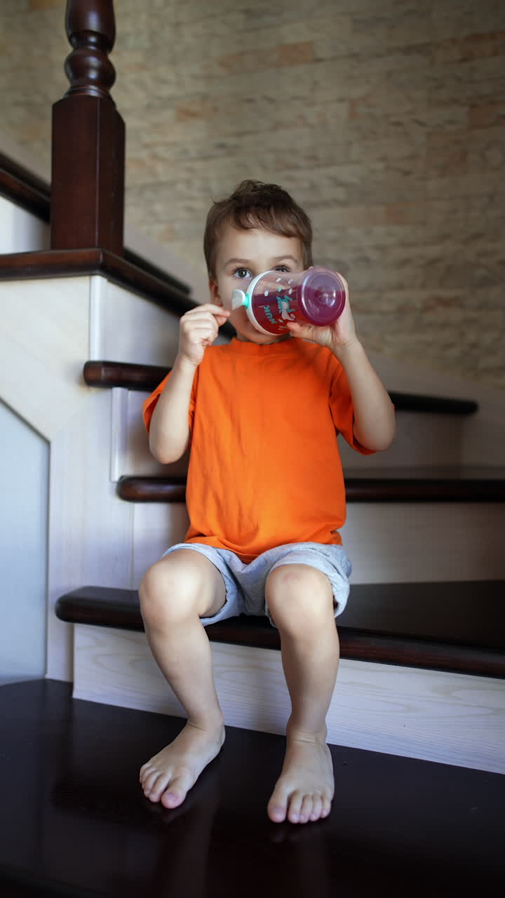 Little kid wearing orange t-shirt sits on the stairs at home. Baby boy drinks juice or compote from a bottle. Vertical video.