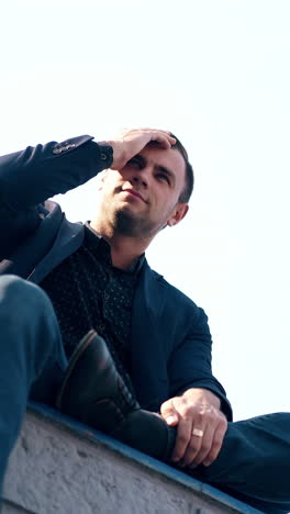 low angle shot guy in suit sits on parapet and fixes black hair by hand with golden ring under white summer sky