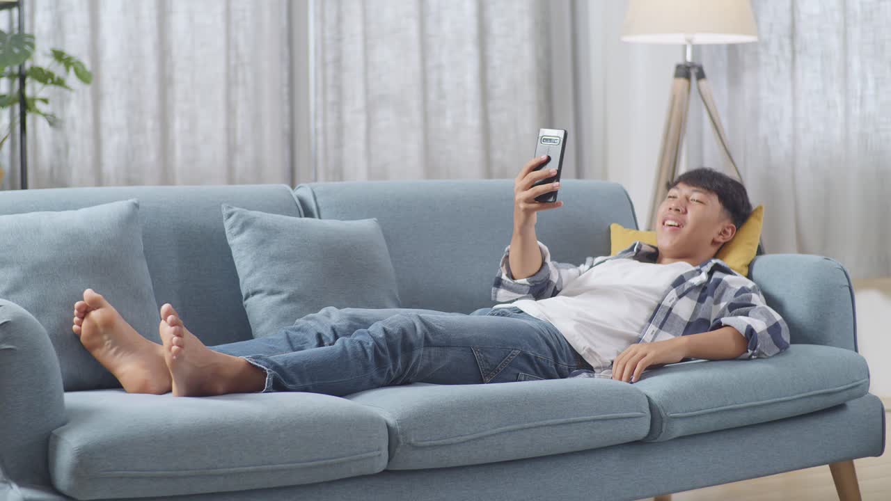 아시안 십대 소년이 집에서 거실의 소파에 누워있는 동안 스마트폰으로 비디오 통화를 하고 있습니다. 손을 흔들고, 미소 짓고, 말하고 있습니다.