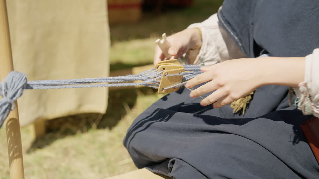 A close-up, handheld shot shows a person in historical costume demonstrating tablet weaving. Their skilled hands manipulate wooden cards to create an intricate pattern in the wool thread