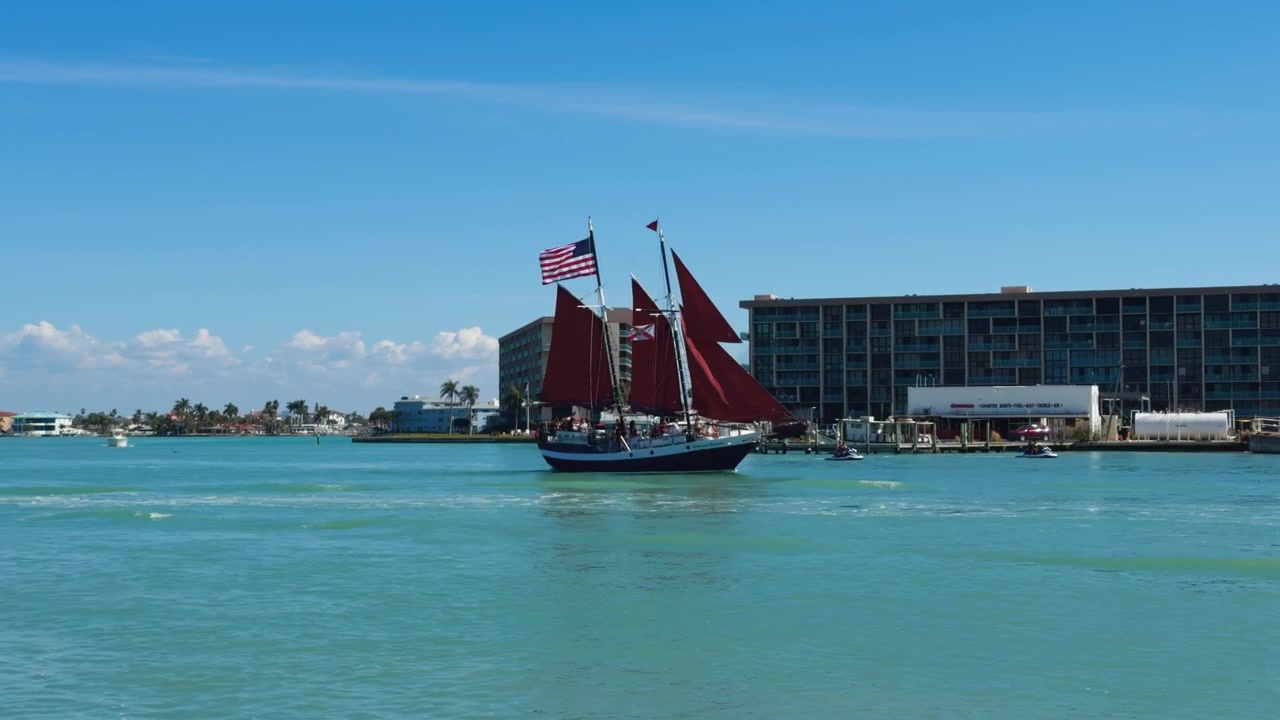 Sidebillede af en smuk sejlbåd med røde sejl og amerikansk flag, der sejler på turkisblåt vand på vej ud på havet foran bygninger ved vandet.