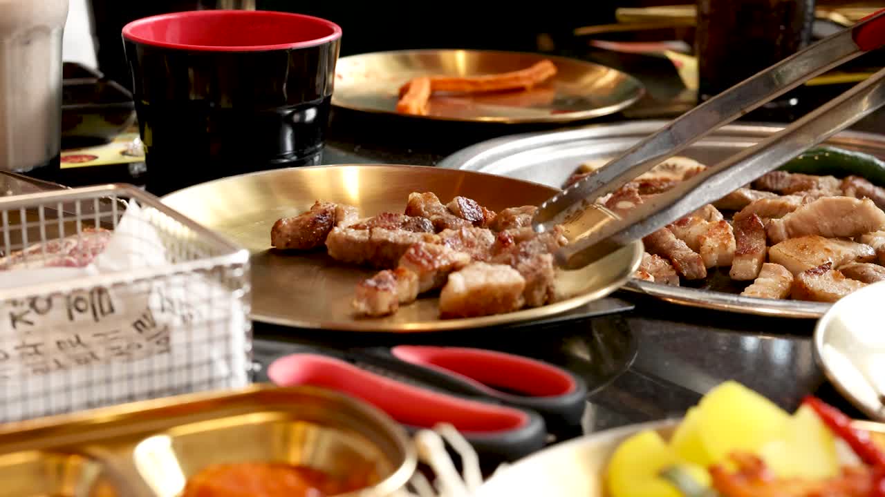 Cooked pork pieces plated with tongs, surrounded by side dishes, under warm indoor lighting