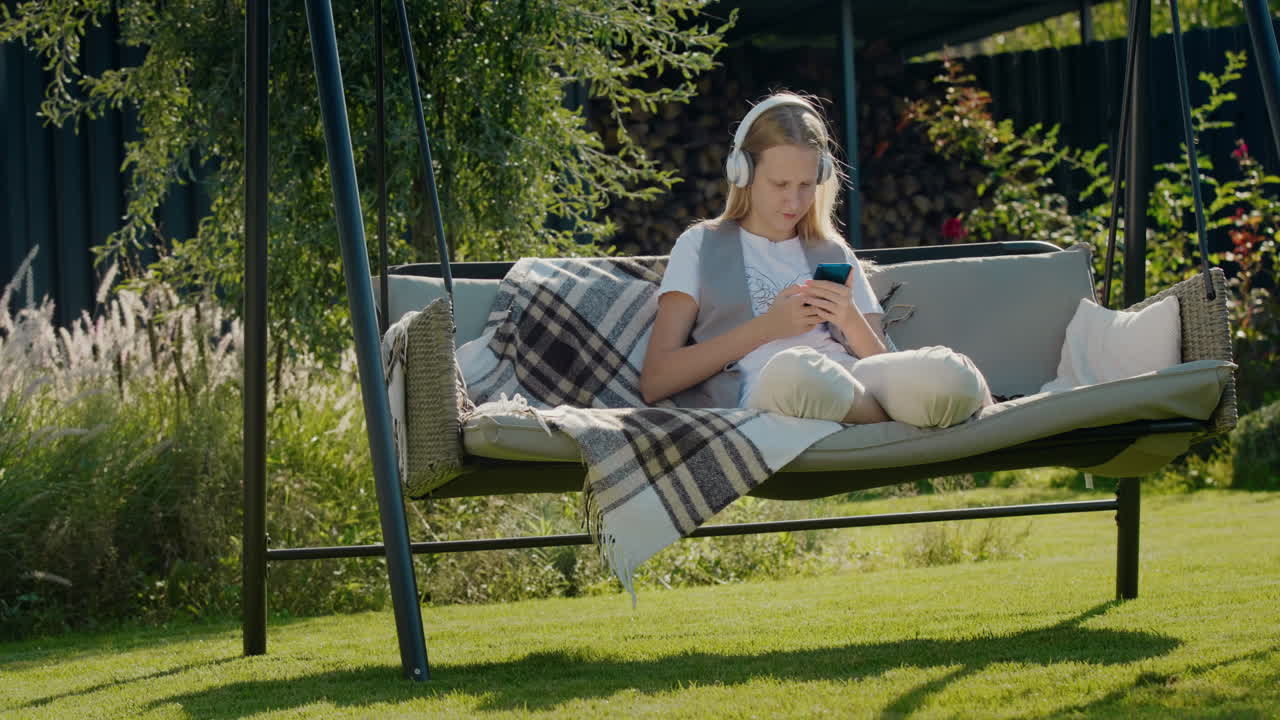 En teenagepige hviler sig på en gyngesofa og bruger en smartphone