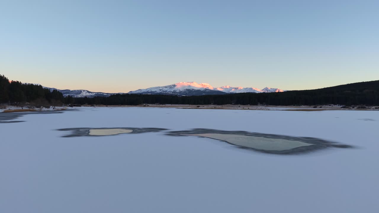 Frozen lake in Esquel, Chubut—Argentina stretches across the foreground, framed by snow-covered peaks and dense forest, drone pushes slowly forward, evoking natural stillness and Patagonian scale
