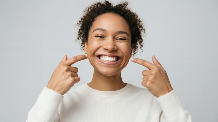 Young woman with curly hair smiling and pointing to her face with both hands in a studio setting