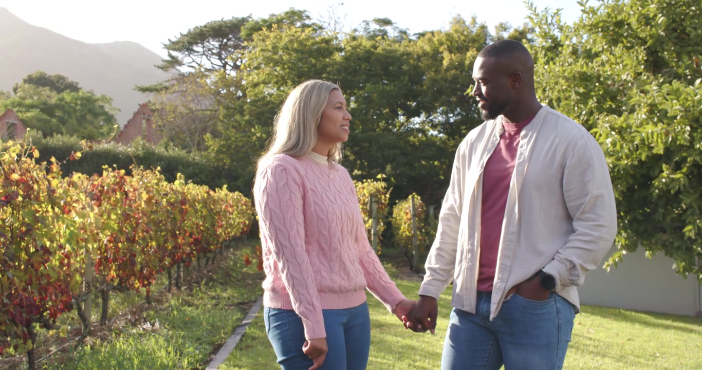 Couple walking along grapevine path, woman speaking while holding smartphone, man listening smiling