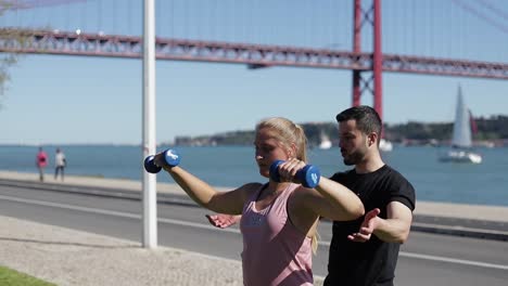 Sportliche-Junge-Frau-Trainiert-Mit-Hanteln-Und-Trainer-Im-Park.