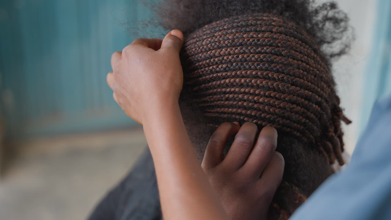 Closeup braided hair being tucked with careful stylist hands, neat cornrows hugging scalp, teal doorway backdrop, patient finishing, precision and texture emphasized, community salon atmosphere