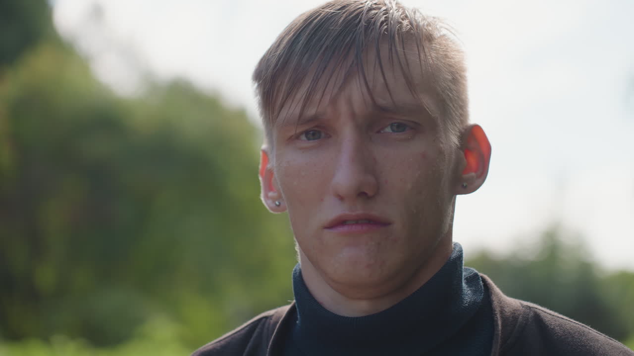 pria basah di luar ruangan setelah hujan, aktor muda menunjukkan perubahan emosional yang halus, tetesan di rambut dan wajah, sweter turtleneck, cahaya alami lembut, shallow depth of field, latar belakang dedaunan hijau yang buram