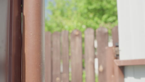 close up of partially open brown wooden gate with bolts in focus as ladies walk through entrance into residential area under clear blue sky with blurred green trees and fence in background