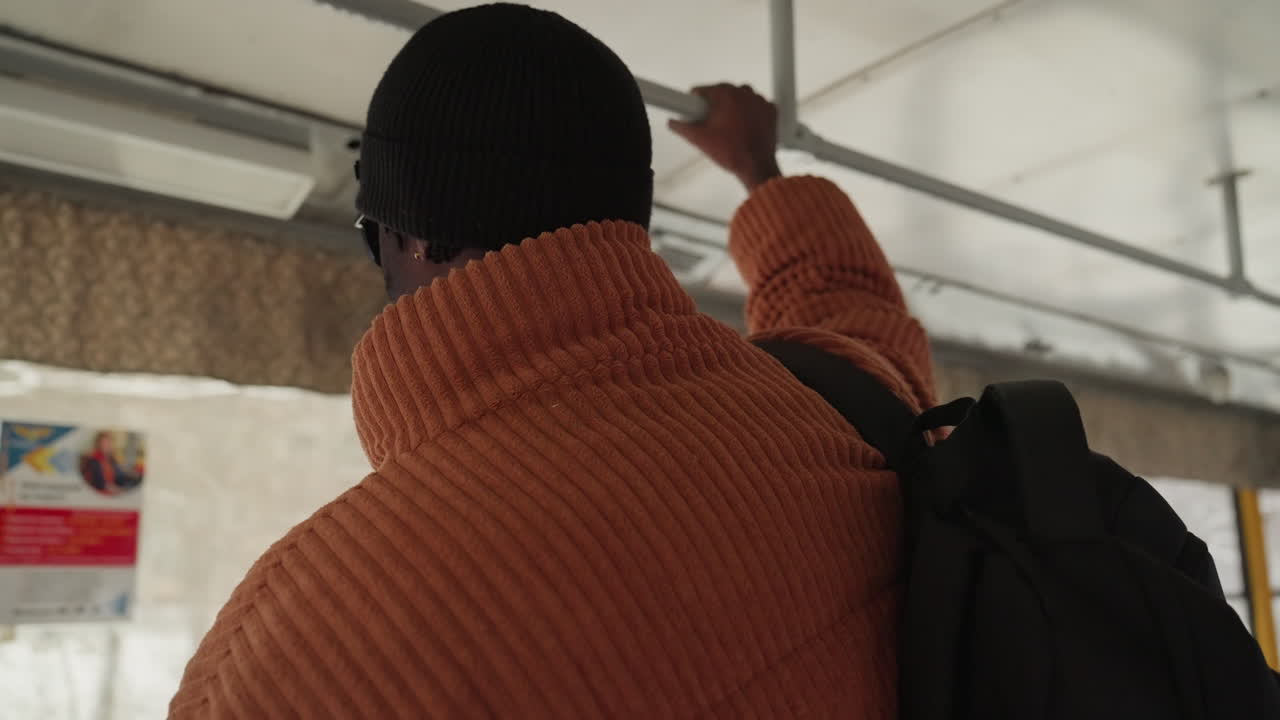 Man back facing bus window wearing chunky orange knit, black beanie, backpack straps visible subtle city posters, muted light, solitude and travel fatigue student returning home after long day